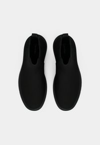 Sapatos pretos de slip-on com uma textura suave, biqueira redonda e painéis laterais elásticos. Apresentam uma aba na parte de trás e uma sola preta plana.