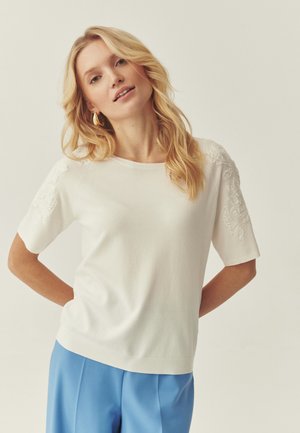 Blondynka o falowanych włosach, ubrana w biały top z krótkim rękawem z haftowanymi rękawami oraz niebieskie spodnie z wysokim stanem na jednolitym tle.