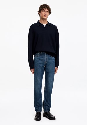 Homme portant un polo noir à manches longues, un jean bleu et des chaussures en cuir noires, debout devant un fond blanc uni.