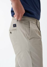 Béžové kalhoty se bočními kapsami, zadní kapsou na knoflík a textilní etiketou. Design je rovný s hladkým povrchem.