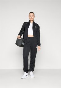 Fato atlético preto com detalhes brancos, apresentando uma jaqueta com fecho e calças com cintura elástica. Inclui uma mala de duffel preta e ténis brancos.