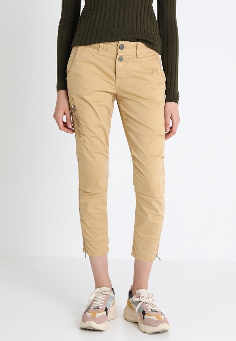 Pantalons cargo beiges avec une coupe ajustée, dotés de deux poches avant, d'une poche latérale zippée et de zips aux chevilles. Portés avec des baskets multicolores.