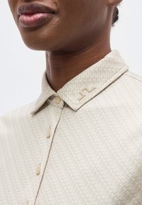 Κοντινό πλάνο ενός ατόμου που φοράει ανοιχτό μπεζ πουκάμισο με μοτίβο και κουμπιά, με μια καρφίτσα για γιακά σε σχήμα γεωμετρικού σχεδίου "JL".