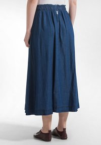 Mujer con una larga falda vaquera azul con cintura elástica y zapatos de cuero burdeos con cordones, de pie sobre un suelo blanco.