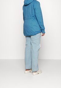 Veste à capuche bleue avec poignets et taille élastiques, associée à un jean bleu clair déchiré et des baskets épaisses de couleur claire.