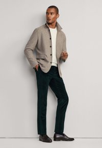 Muž v béžovej sako so zapínaním na gombíky cez biely sveter, tmavozelené nohavice a tmavohnedé mokasíny, stojaci pred jednofarebnou šedou stenou.