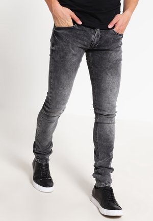 Homme portant un jean slim gris délavé avec des baskets noires, les mains dans les poches, debout sur un fond blanc.