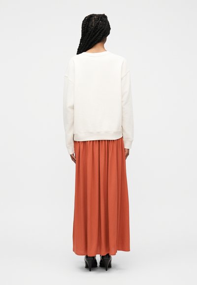 Γυναίκα με μακριά πλεκτή κοτσίδα που φοράει λευκό φούτερ και μακριά φούστα σε απόχρωση σκουριάς πορτοκαλί, στέκεται γυρισμένη με την πλάτη προς τα πίσω σε απλό φόντο.