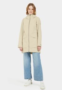 Veste à capuche beige clair avec une fermeture éclair sur le devant, deux grandes poches et une coupe droite. Associée à un jean bleu à jambes larges et des baskets blanches.
