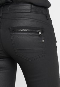 Pantalones vaqueros skinny negros con un acabado brillante, que cuentan con un bolsillo trasero con cremallera y herrajes plateados, además de un sutil logo en la cinturilla.