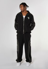 Svart träningsställ med vita pipedetaljer; jacka med dragkedja och matchande byxor. Figursydd och avslappnad design med logotypdetaljer. Vita sneakers.