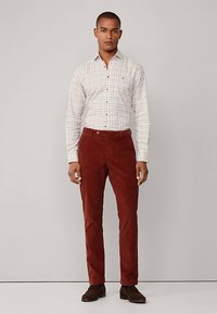 Camisa blanca a cuadros con líneas rojas y verdes, combinada con pantalones de pana color óxido. Calzado marrón completa el conjunto, posando de pie.