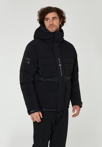 Svart puffjacka med hög krage, justerbar huva, flera framfickor och en slimmad design. Har kontrastpaneler och varumärkeslogotyp.