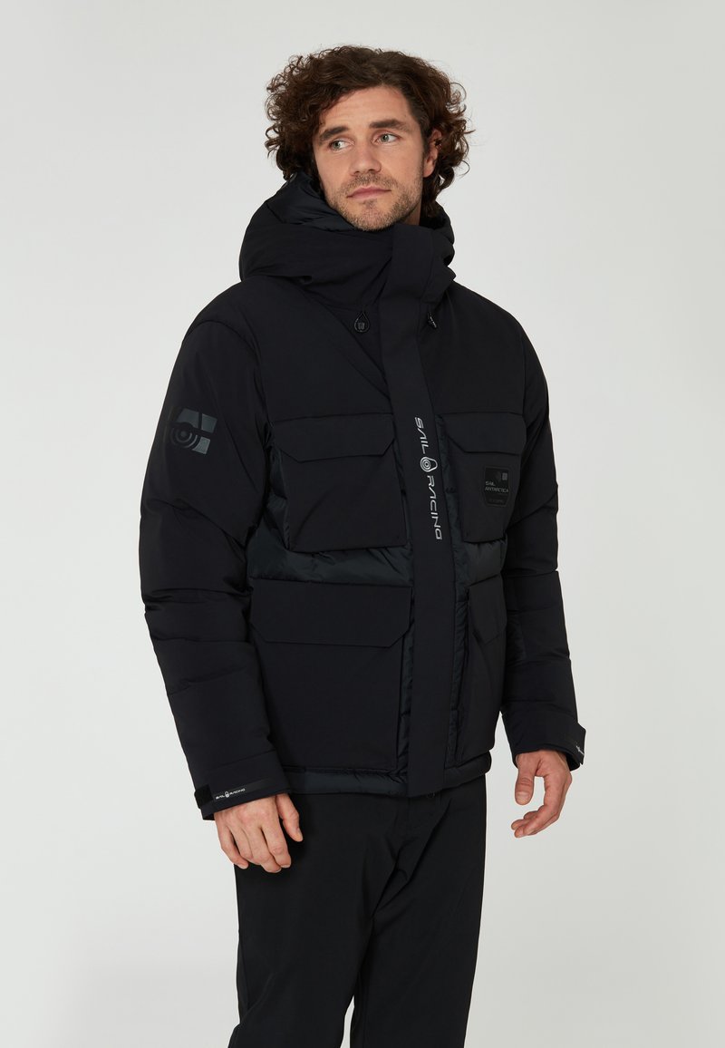 Svart puffjacka med hög krage, justerbar huva, flera framfickor och en slimmad design. Har kontrastpaneler och varumärkeslogotyp.