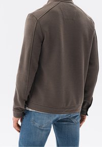 Mężczyzna w teksturowanej brązowej kurtce i niebieskich dżinsach, pokazany od tyłu na jednolitym tle.