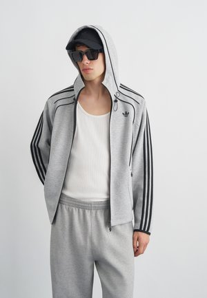 Młody mężczyzna ubrany w szary dres Adidas z czarnymi paskami, białą koszulkę bez rękawów, czarną czapkę i duże czarne okulary przeciwsłoneczne na jednolitym tle.