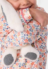 Un tissu à motif floral dans des couleurs pastel présente un col doux et moelleux ainsi que des accents en forme de lapin avec des détails rayés sur les pattes.