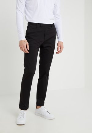 Mężczyzna ubrany w czarne spodnie o dopasowanym kroju, białą koszulę z długim rękawem i białe trampki, stojący na tle jednolitego jasnego tła.