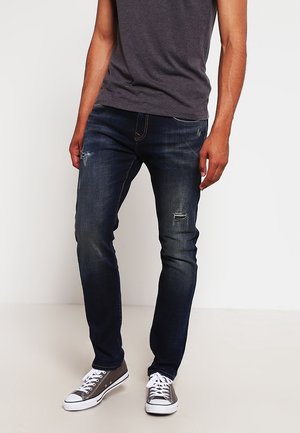 Homme portant un jean bleu foncé déchiré, coupe slim, et des baskets basses grises, debout sur un fond blanc.