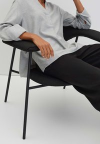 Personne portant un haut gris clair à manches longues et un pantalon noir, assise sur une chaise moderne noire avec accoudoirs, un bras reposant sur la chaise.