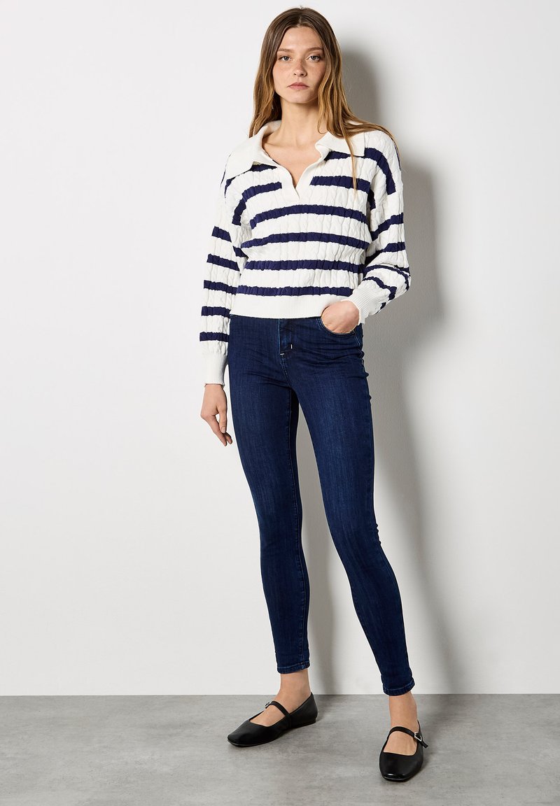 Pull rayé blanc et marine avec col, tissu texturé, associé à un jean skinny bleu foncé et des chaussures plates noires.