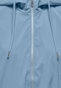 Svetlo modra jopa s kapuco, s srebrnim zadrgo na sredini in usklajenimi vrvicami z metalnimi konicami na obeh straneh.