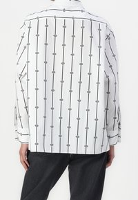 Chemise blanche à manches longues avec un motif de lignes verticales noires et des accents géométriques. Le tissu semble lisse et léger.