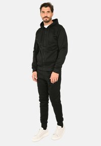 Felpa nera con zip e cappuccio regolabile, abbinata a pantaloni jogger neri e sneakers bianche. Tessuto liscio e casual.