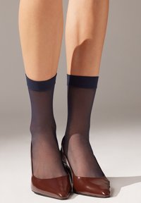 Calzini alla caviglia di nylon trasparente blu scuro con bordo a coste di un blu notte testurizzato, indossati con scarpe con tacco a punta in pelle verniciata marrone. Gambe visibili dalla metà del polpaccio in giù.