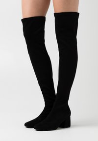 Botas negras de ante hasta la rodilla con punta afilada y tacón grueso. Textura suave con un ajuste ceñido alrededor de la pierna.