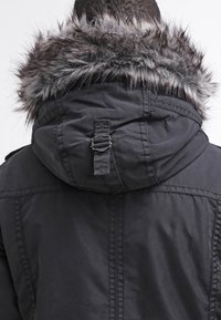 Giacca parka nera con cappuccio rimovibile foderato in pelliccia sintetica, dettagli con cinghie regolabili e un tessuto liscio e strutturato.