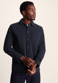 Navy corduroy shirt featuring a standard collar, front buttons, a chest pocket, and long sleeves with buttoned cuffs. Textured fabric.