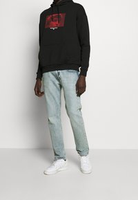 Svart huvtröja med röd grafisk tryck och vit text, kombinerad med ljusblå jeans och vita sneakers.