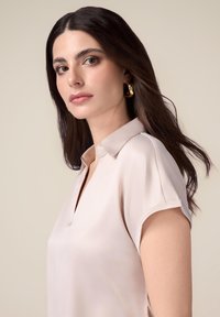Lichtroze blouse met een zachte glans, voorzien van een V-hals en korte opgerolde mouwen. Model draagt gouden hoopoorbellen en heeft steil, donker haar.