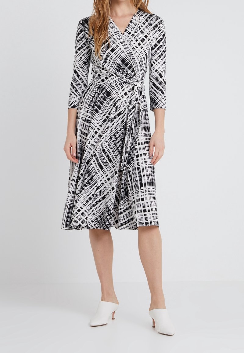 Femme portant une robe portefeuille à carreaux noir et blanc avec des manches trois-quarts et des mules blanches à talons, debout devant un fond uni.