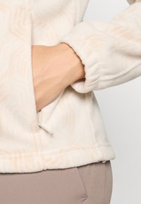 Chaqueta de forro polar en color blanco roto con un sutil patrón ondulado, puños elásticos y un bolsillo lateral. Textura suave y diseño ligero.