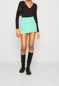Falda mini de pana verde menta con botones en el frente, combinada con una blusa negra de manga larga ajustada y botas negras hasta la rodilla.