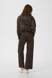 Moteris su raudonais plaukais, vilkinti tamsiai rudos odos striukę ir derančias plačias kelnes, dėvi baltus, smailėjančius aukštakulnius, stovinti nugara.