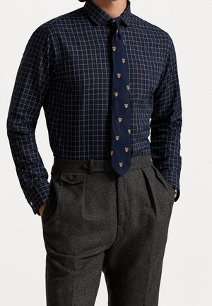 Mężczyzna w granatowej koszuli w kratę z granatowym krawatem ozdobionym małymi emblematami tarcz, wpuszczoną w wysokowalne, szare spodnie z plisami.