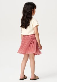 Geel T-shirt met korte mouwen en een roze plisségordelrok, met een gerimpelde tailleband, gecombineerd met beige instappers, gezien van achteren.