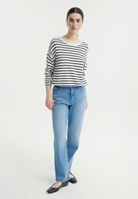 Gestreepte, lang mouwtop in wit en marineblauw, gecombineerd met lichtblauwe rechte jeans. Model draagt zwarte ballerina's en staat op een witte achtergrond.