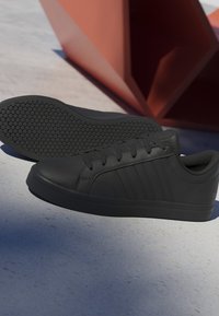 Paire de baskets basses noires avec semelle texturée, posées sur un béton clair près d'une sculpture géométrique rouge qui projette des ombres.