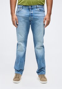 Άνδρας που φοράει ανοιχτό γαλάζιο τζιν παντελόνι, πουκάμισο ελιάς και μπεζ αθλητικά παπούτσια, στέκεται μπροστά σε απλό λευκό φόντο.