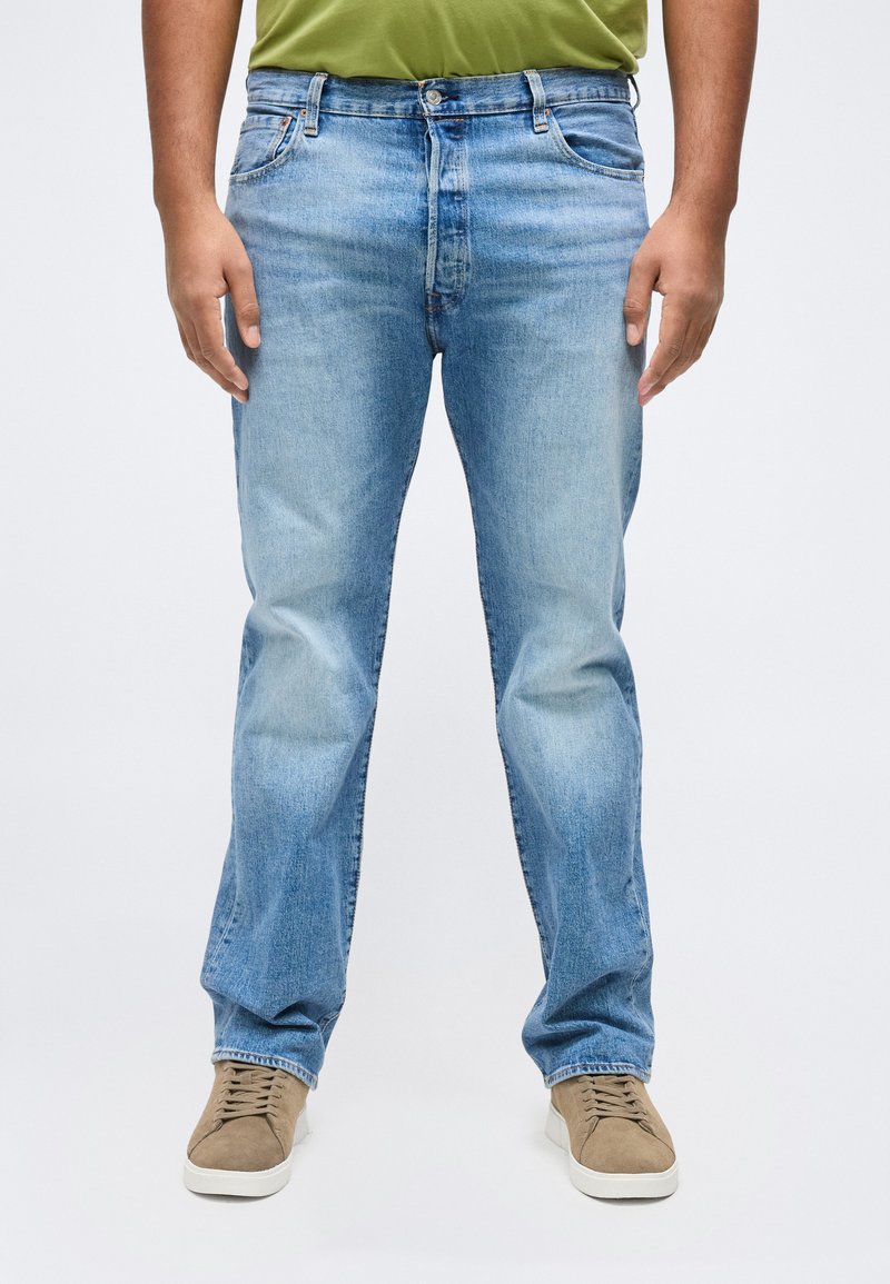 Άνδρας που φοράει ανοιχτό γαλάζιο τζιν παντελόνι, πουκάμισο ελιάς και μπεζ αθλητικά παπούτσια, στέκεται μπροστά σε απλό λευκό φόντο.