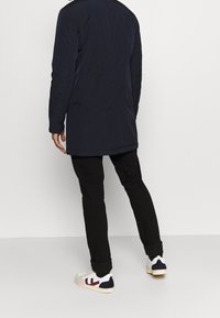 Marineblauer langer Mantel mit glatter Textur, kombiniert mit schwarzen Hosen und weißen Sneakers mit weinroten und marineblauen Akzenten. Minimalistisches Design.