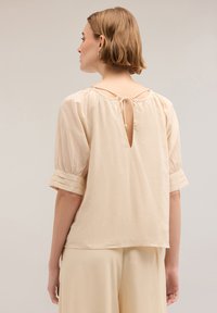 Beige blouse met korte pofmouwen en een sleutelgatdetail op de rug, vastgebonden met een touwtje, met subtiele plooien op de manchet van de mouwen.