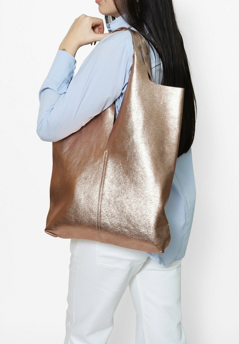 Sac fourre-tout en or rose métallique avec une texture lisse, de forme oversize et un détail de fermeture éclair unique. Porté sur l'épaule avec une chemise bleu clair.