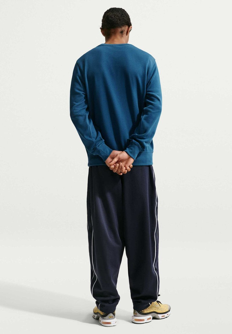 Felpa a maniche lunghe color verde petrolio, pantaloni blu navy con strisce laterali bianche e scarpe da ginnastica multicolori. Il tessuto sembra morbido con una vestibilità comoda.