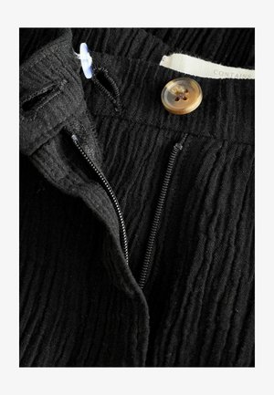 Mustra musta tekstuuriga riie, millel on nähtav tõmblukk ja beež nupp sulgemiseks. Omab horisontaalseid voldikesi ja sildit, millel on märge "sisaldab".