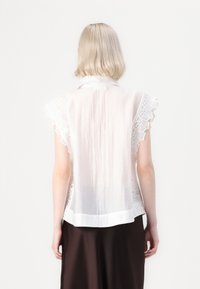 Camicia bianca con bordi in pizzo lungo le maniche a sbuffo e l'orlo inferiore, realizzata in un tessuto leggero e trasparente, con colletto arrotondato.